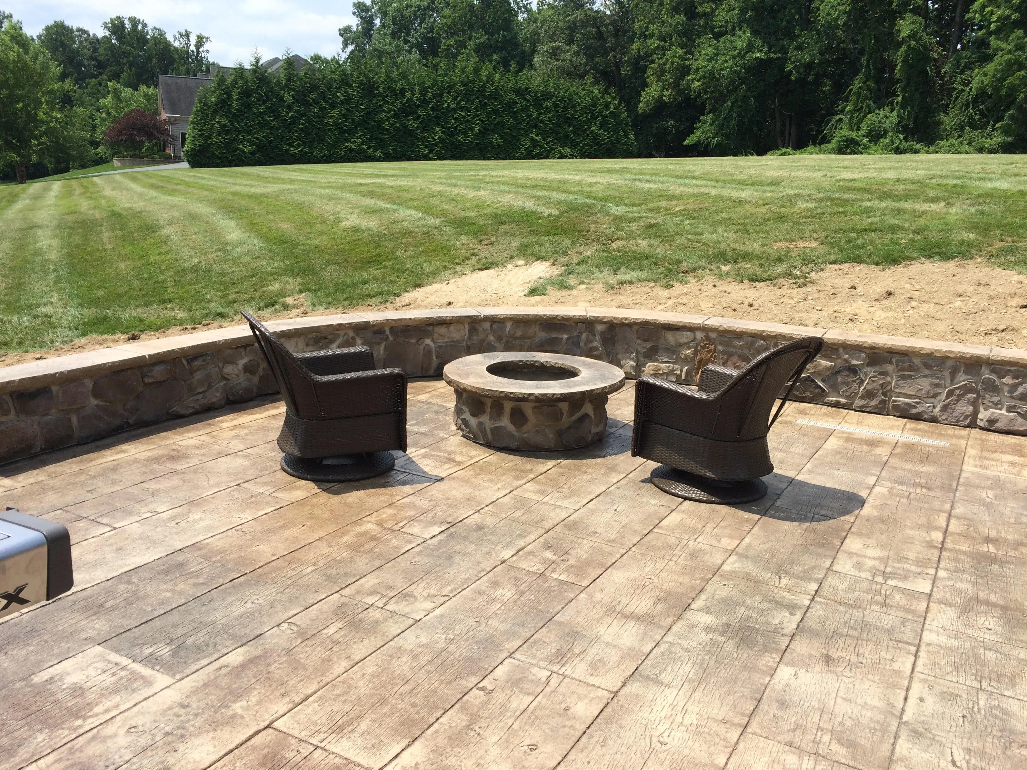 Stamped concrete patio with stone seating wall, fire pit, and wicker chairs overlooking lawn