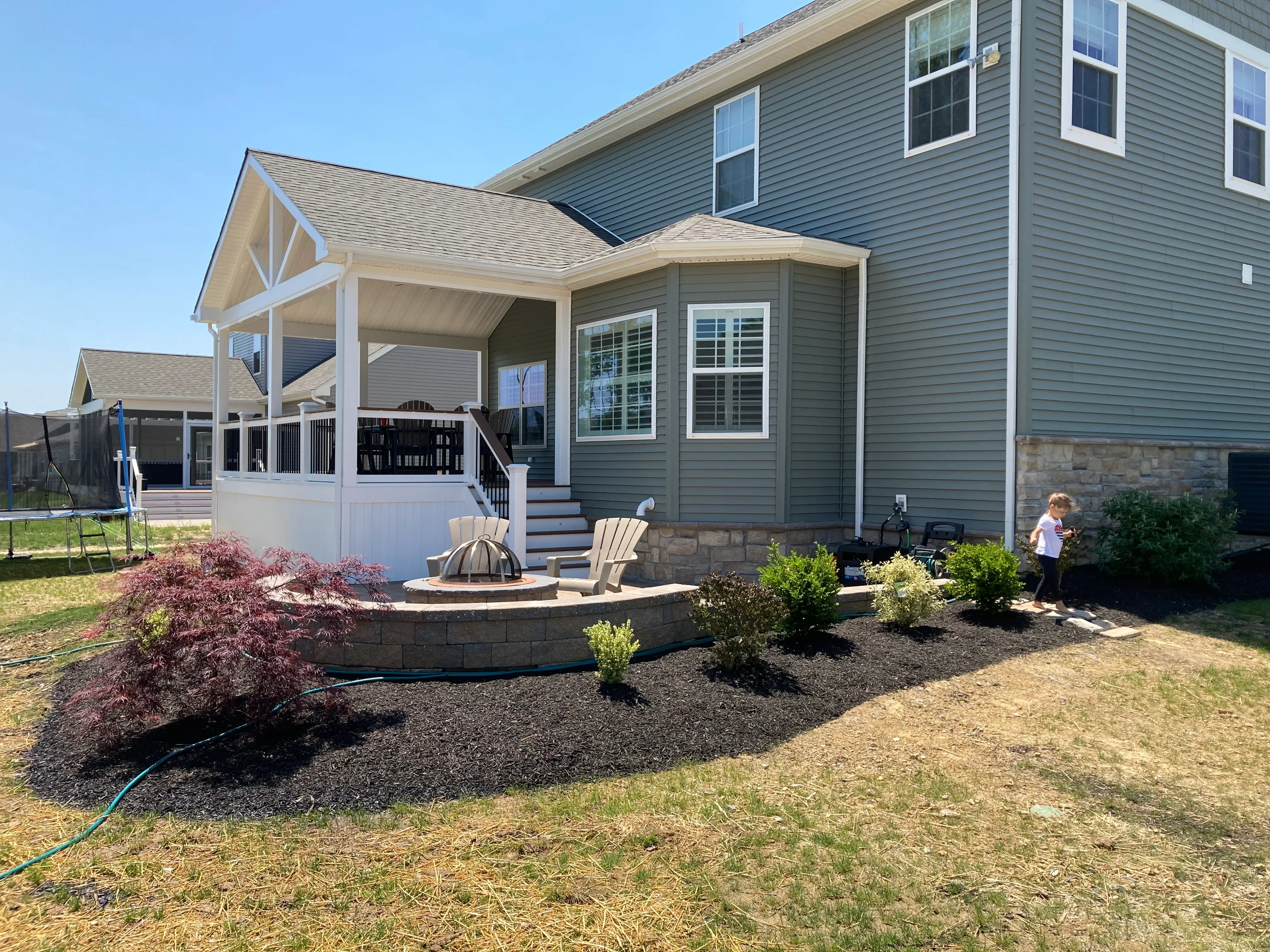 White covered patio with vinyl railings, stone fire pit, and landscaped mulch beds with shrubs