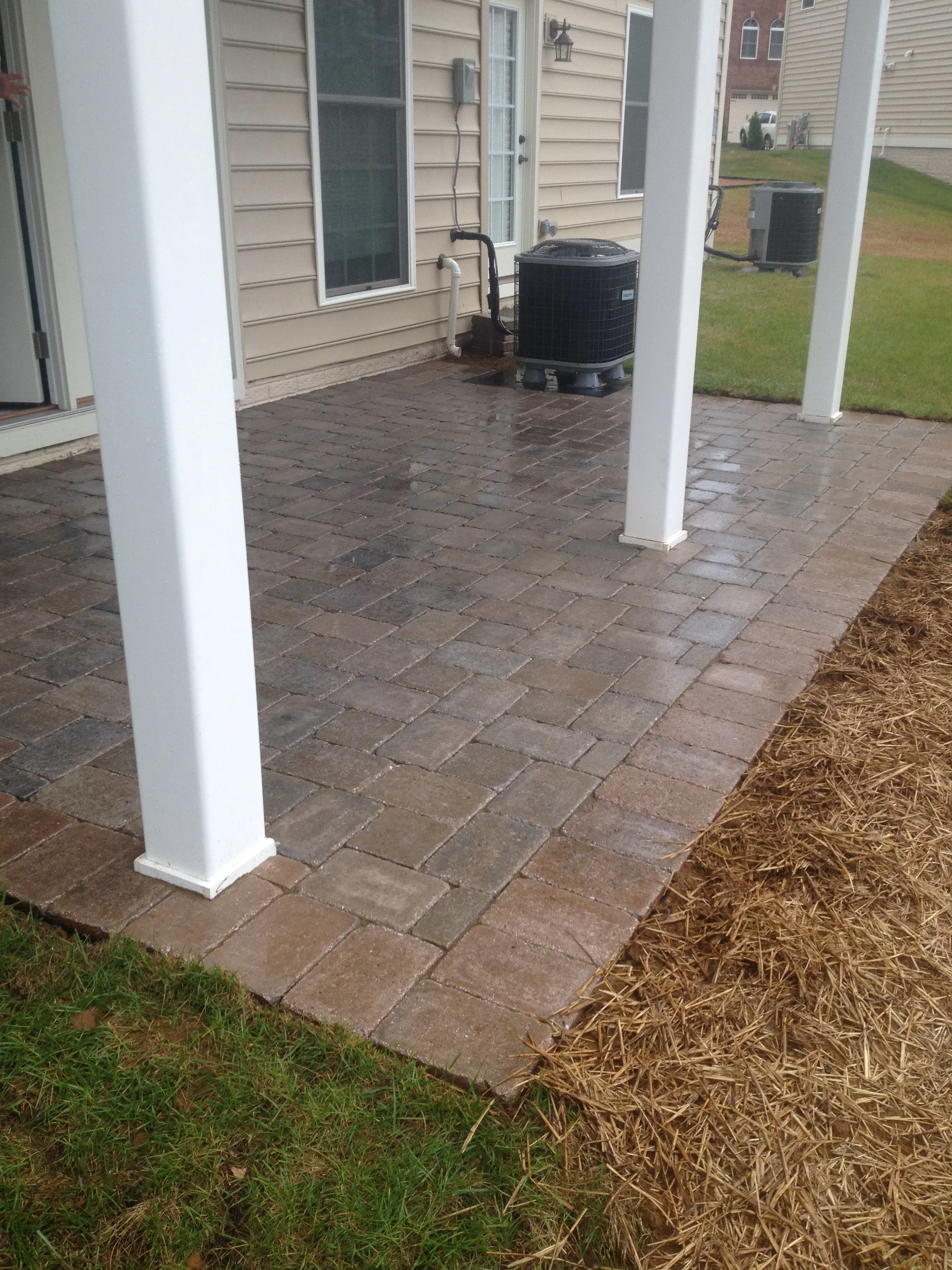 Brown paver patio with white columns under covered structure, adjacent to house siding